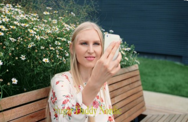 Sieviete puķu cepurē veic videozvanu parkā, sēžot uz soliņa.