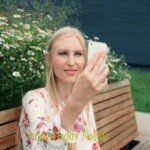 Kukkapuseroinen nainen soittaa videopuhelua puistossa istuen penkillä.